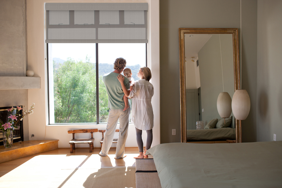 Couple holding a child in a bedroom with motorized shades partially lowered over large windows offering outdoor views.