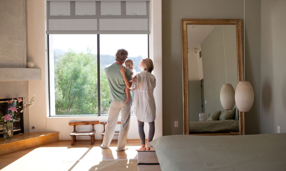 <span class="uk-h4">Couple holding a child in a bedroom with motorized shades partially lowered over large windows offering outdoor views.</span>